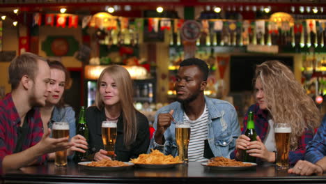 Multiethnische-Grappa-Freunde-Sitzen-In-Einer-Bar,-Trinken-Bier,-Unterhalten-Sich-Amüsant-Und-Essen-Chips.